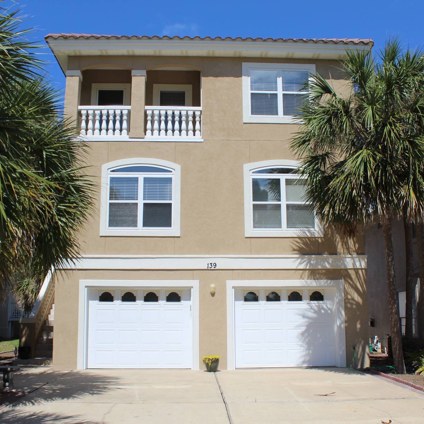 Three-story coastal home with tan stucco, white trim, balconies, and palm trees